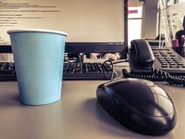 taza de café azul de cartón de una cafetería en un escritorio con una computadora con teclado y mouse y suministros de oficina de un trabajador de oficina durante el almuerzo. pausa para el café de negocios foto