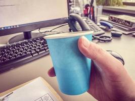 taza de café azul de cartón de una cafetería en un escritorio con una computadora con teclado y mouse y suministros de oficina de un trabajador de oficina durante el almuerzo. pausa para el café de negocios foto