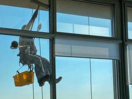 un trabajador de lavado de ventanas masculino, escalador industrial cuelga de un edificio alto, rascacielos y lava grandes ventanas de vidrio para limpieza en lo alto de una gran ciudad foto