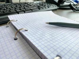 A writing pen rests on a notepad with squared paper sheets on a work desk with stationery in a business office photo