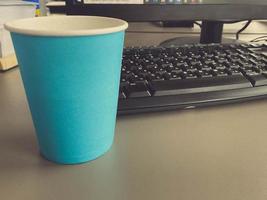 Cardboard blue coffee cup from a coffee shop on a desktop with a computer with a keyboard and mouse and office supplies of an office worker during lunch. Business coffee break photo