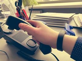 A man's hand in a shirt holds a landline phone receiver on a work desk with office supplies in a business office photo