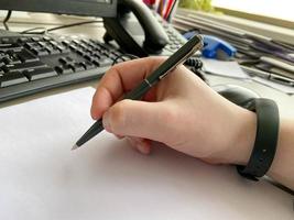 A man's hand in a shirt and with a fitness bracelet holds a pen and writes on the table at the office table with a computer with a keyboard. Business work photo
