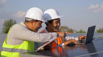 el ingeniero inspector asiático tiene un tablero de control y un joven tecnológico usa una computadora portátil, dos ingenieros están discutiendo durante el trabajo en la granja solar, la estación de paneles solares en segundo plano video