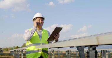 Slow motion shot, Young engineer in white helmet holding checking board in hand standing and talking on smartphone while working at solar farm in background video