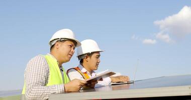 asiatischer Inspektor-Ingenieurmann, der ein Kontrollbrett hält, und junger Fachmann verwenden Laptop-Computer, zwei Ingenieure diskutieren während der Arbeit im Solarpark, Solarpanel-Station im Hintergrund video