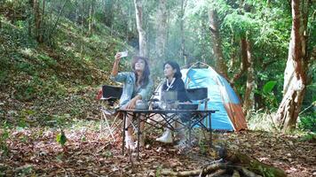 Handheld shot, front view Young Asian pretty woman and her girlfriend sitting at front of tent, use mobile phone take photo during camping in forest with happiness together video