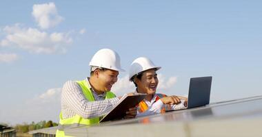 ingénieur inspecteur asiatique homme tenant un tableau de contrôle et jeune homme spécialiste utilisant un ordinateur portable, deux ingénieurs discutant et heureux de réussir pendant le travail à la ferme solaire, panneau solaire en arrière-plan video