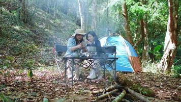 handhållen skott, främre se ung asiatisk Söt kvinna och henne flickvän Sammanträde på främre av tält, använda sig av mobil telefon ta Foto under camping i skog med lycka tillsammans video