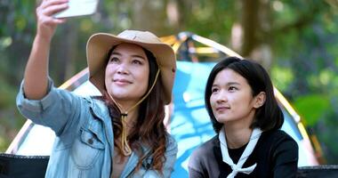 Handheld shot, front view Young Asian pretty woman and her girlfriend sitting at front of tent, use mobile phone take photo during camping in forest with happiness together video