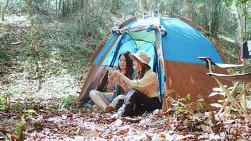 Handheld shot, front view Young Asian pretty woman and her girlfriend sitting at front of tent, use mobile phone take photo, talking and show picture during camping in forest with happiness together video