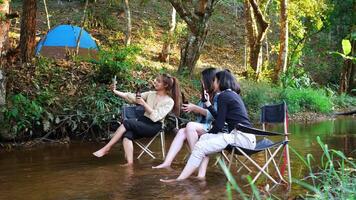 toma manual, un grupo de jóvenes asiáticas beben cerveza en sus sillas y se mojan los pies en el arroyo mientras acampan en el parque natural, disfrutan hablando y riéndose juntas. video