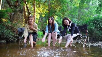 tiro portátil, grupo de jovens mulheres asiáticas sentadas em suas cadeiras e gostam de espirrar água no riacho enquanto acampam no parque natural, elas estão conversando e rindo divertidas juntas. video