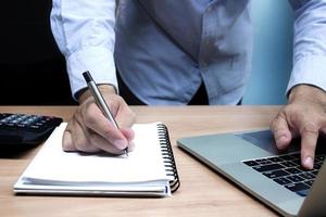 mano de hombre de negocios sosteniendo un bolígrafo para papeleo con una computadora portátil en la mesa foto