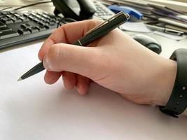 A man's hand in a shirt and with a fitness bracelet holds a pen and writes on the table at the office table with a computer with a keyboard. Business work photo