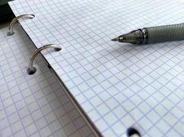 A writing pen rests on a notepad with squared paper sheets on a work desk with stationery in a business office photo