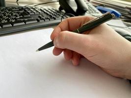 A man's hand in a shirt and with a fitness bracelet holds a pen and writes on the table at the office table with a computer with a keyboard. Business work photo