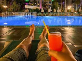 piernas de hombres en una tumbona junto a la piscina por la noche, relajándose con un cóctel alcohólico en un hotel en un cálido paraíso tropical del este del país foto