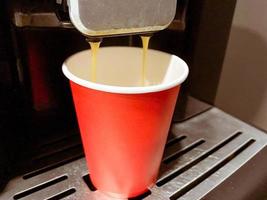 Red cardboard coffee cup into which delicious invigorating hot black Americano coffee is poured from the coffee machine in the office in the morning photo