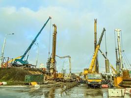 equipo de construcción en el sitio de reparación del paso elevado. las grúas levantan grandes bloques de hormigón. junto a un camión grande. sitio de construcción bajo la lluvia con charcos foto