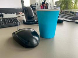 taza de café azul de cartón de una cafetería en un escritorio con una computadora con teclado y mouse y suministros de oficina de un trabajador de oficina durante el almuerzo. pausa para el café de negocios foto