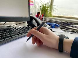 A man's hand in a shirt and with a fitness bracelet holds a pen and writes on the table at the office table with a computer with a keyboard. Business work photo