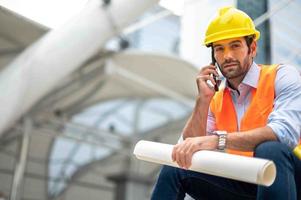 el ingeniero caucásico usa un teléfono inteligente para hablar, usa chaleco naranja y un gran sombrero duro, y la otra mano sostiene el plano de planta blanco en el sitio de trabajo del centro de la ciudad. foto