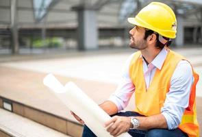 ingeniero caucásico con chaleco naranja y sombrero duro grande, y la otra mano sosteniendo el plano de planta blanco en el sitio de trabajo del centro de la ciudad. foto