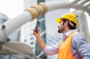 Caucasian man engineer use a smartphone for talking, wearing orange vest and big hard hat, and the other hand holding the white floor plan in the site work of the center city.