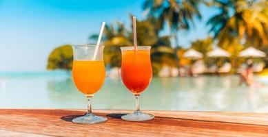 Cocktail glasses at summer poolside, resort beach side. Tropical island closeup outdoor bar cafe or restaurant with wooden table two cocktails, blurred umbrellas beds and calm swimming pool recreation photo