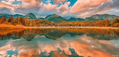 Colorful autumn sunrise clouds sky over lake. Panoramic nature landscape, peaceful dream water surface, sunset autumnal background. Forest lake, mountains around. Peaceful amazing nature panorama