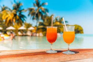 copas de cóctel en la piscina de verano, junto a la playa del complejo. isla tropical, primer plano, bar, cafetería al aire libre o restaurante con mesa de madera, dos cócteles, sombrillas borrosas, camas y recreación tranquila en la piscina foto