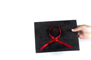 Hand holding a red ribbon pasted on a blackboard signifying concern for people with HIV aids isolated on a white background photo