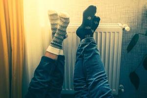 Couple hold legs up heating feet in cold home indoors on radiator in winter with cozy winter stylish woolen socks on. Valentines funny together warm feet by radiator by window
