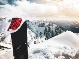 Black Snowboard with christmas hat stand in snow beautiful white caucasus mountains in Gudauri ski resort top viewpoint in winter. Georgia skiing winter holiday destination photo