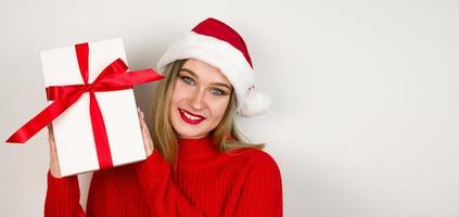 Closeup portrait of blonde girl wearing santa hat,red cozy sweater and holding gift in her hands.Banner with place for text photo