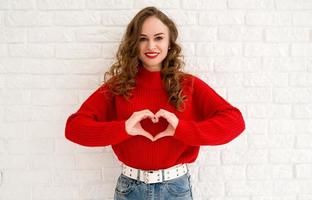 niña sonriente en suéter rojo que muestra el corazón con dos manos, signo de amor. aislado contra la pared blanca. día de la mujer tarjeta de san valentín foto