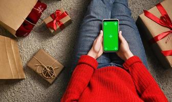 Top view on Unrecognizable woman holding phone with green screen surrounded with gifts. Christmas shopping or social media concept. photo