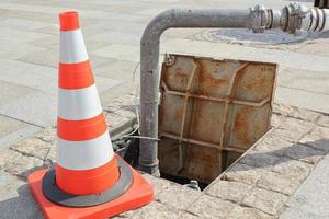 Open manhole, construction traffic warning cone and metal pipe on sidewalk. Concept of repair of supply system, underground utilities, cable laying, water accident. Troubleshooting cleaning sewage. photo