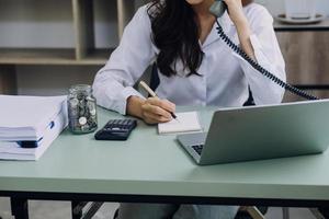 mujer de negocios joven sentada en la oficina en la mesa y usando un teléfono inteligente. en el escritorio hay una computadora portátil y una tableta, en gráficos y gráficos en pantalla. mujer analizando datos. estudiante aprendiendo en línea. foto