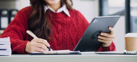 Young business woman sitting in office at table and using smartphone. On desk is laptop and tablet computer, on screen charts and graphs. Woman analyzing data. Student learning online. photo