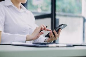 Young business woman sitting in office at table and using smartphone. On desk is laptop and tablet computer, on screen charts and graphs. Woman analyzing data. Student learning online. photo