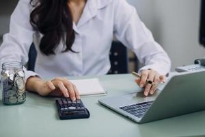 Young business woman sitting in office at table and using smartphone. On desk is laptop and tablet computer, on screen charts and graphs. Woman analyzing data. Student learning online. photo