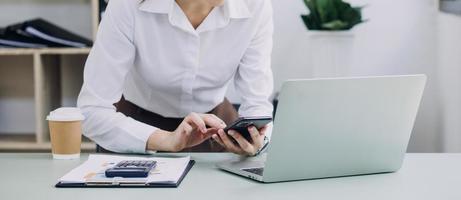 Young business woman sitting in office at table and using smartphone. On desk is laptop and tablet computer, on screen charts and graphs. Woman analyzing data. Student learning online. photo