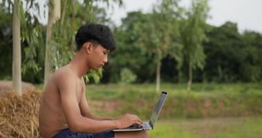 un joven granjero sin camisa se sienta en el suelo, escribe en una computadora portátil y mira los campos de arroz