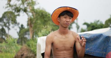 de cerca, el joven granjero en topless sentado junto al pajar se limpió el sudor de la frente y se limpió el calor con un sombrero en el campo de arroz, se siente cansado video