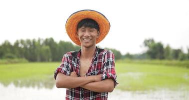 vista frontal, primer plano de un agricultor, retrato de un adulto joven con camisa a cuadros y sombrero de pie con los brazos cruzados, ríe y mira a la cámara con timidez. campo de arroz en el fondo video