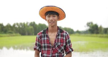 vista frontal, primer plano de un agricultor, retrato de un adulto joven con camisa a cuadros y sombrero de pie con los brazos cruzados, sonriendo y mirando a la cámara. campo de arroz en el fondo video