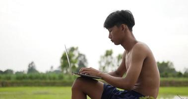 un joven granjero sin camisa se sienta en el suelo, escribe en una computadora portátil y mira los campos de arroz