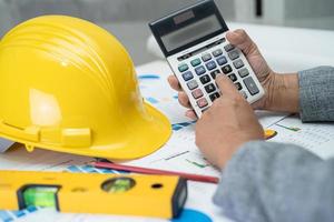 Asian engineer with architectural house plan project blueprint, graph and yellow helmet and engineering construction tools. photo